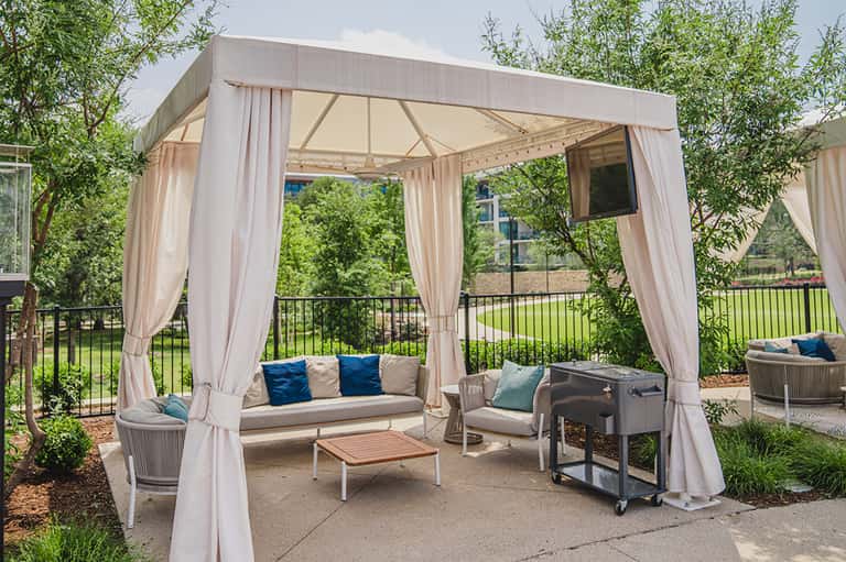 Outdoor lounge pavilion with cream curtains, seating, coffee table, and lush green surroundings