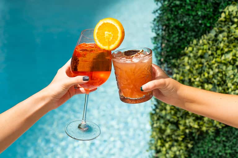 Two cocktails clinking poolside with orange slice garnish and tropical backdrop
