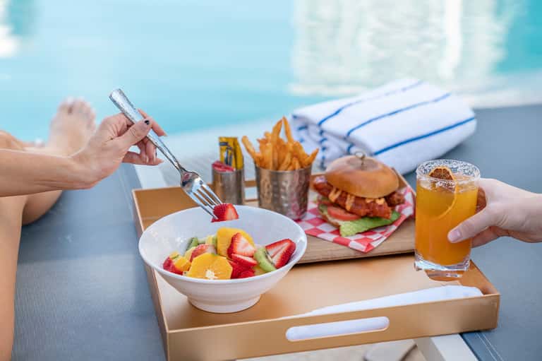 Beach poolside meal with fruit bowl, burger, fries, and cold beverage service