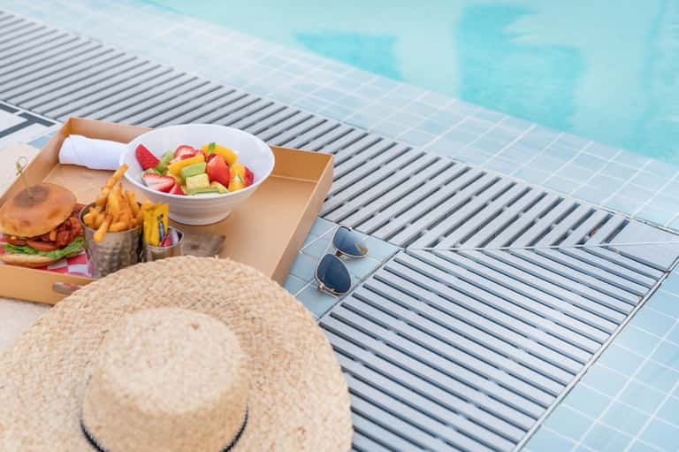 Poolside lunch setup with sandwich, fruit, and sunglasses on wooden deck next to pool