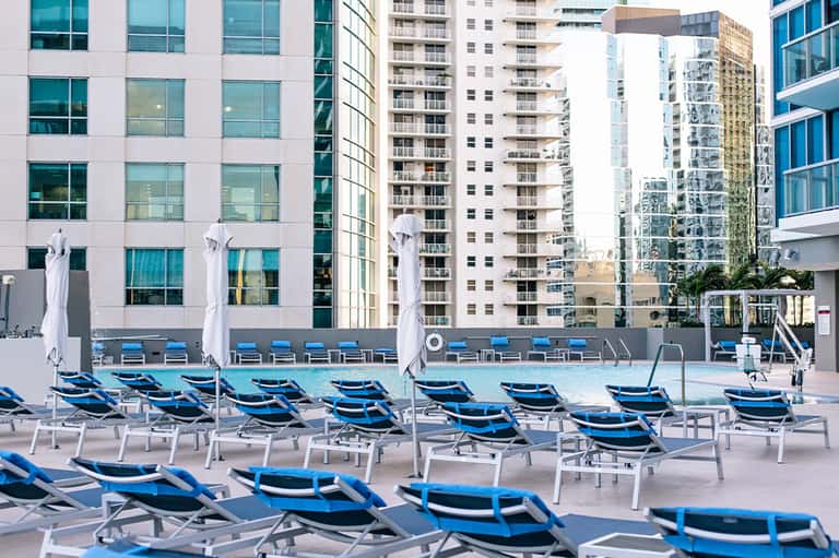 Urban rooftop pool with blue lounge chairs, white umbrellas, and high-rise residential buildings