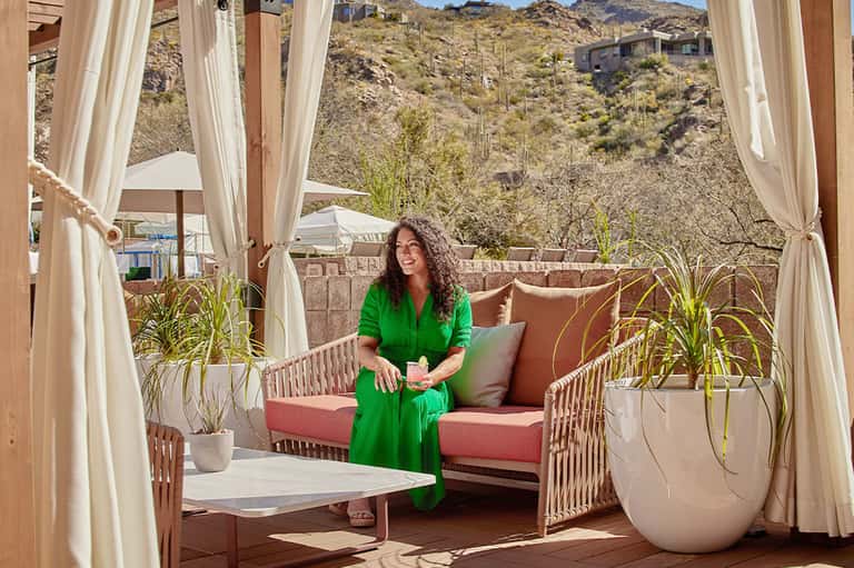 Woman in green dress sitting on coral cushioned seating with desert mountain views and curtains
