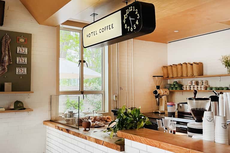 Hotel coffee station with vintage clock sign, wooden counter, shelving with supplies, and green plants