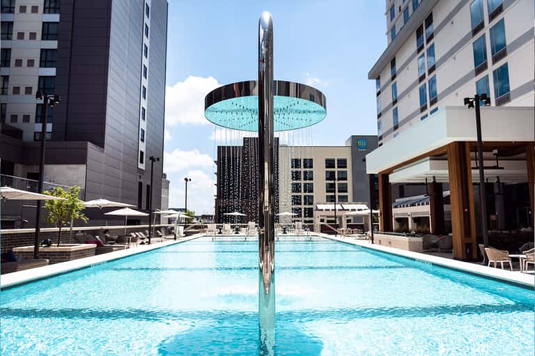 Modern urban pool with cylindrical rain shower sculpture between two turquoise lap pools and tall buildings