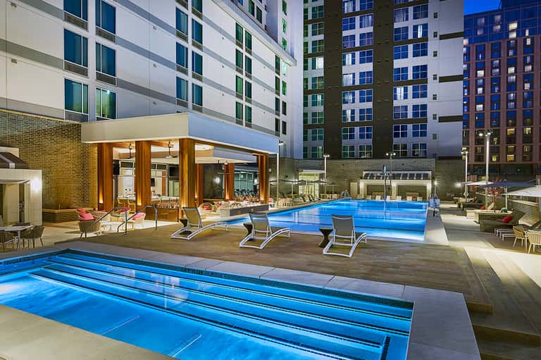 Urban resort pool area at night with illuminated lap pool and loungers near modern buildings