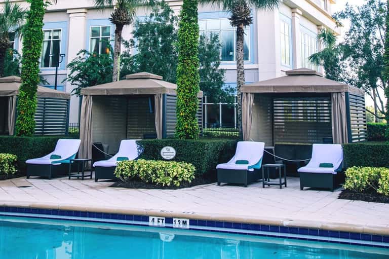 Modern poolside seating area with white loungers, dark pergolas, ivy-covered walls, and manicured hedges