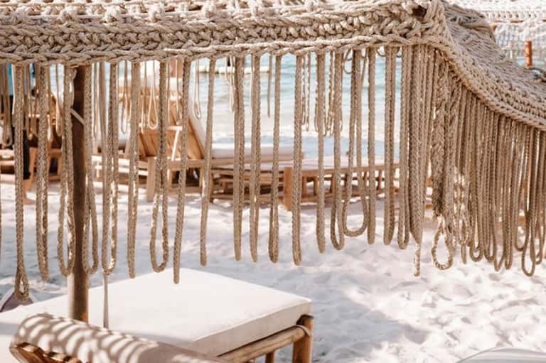 Beachside cabana with macramé fringe canopy, white cushioned seating, and ocean view through ropes