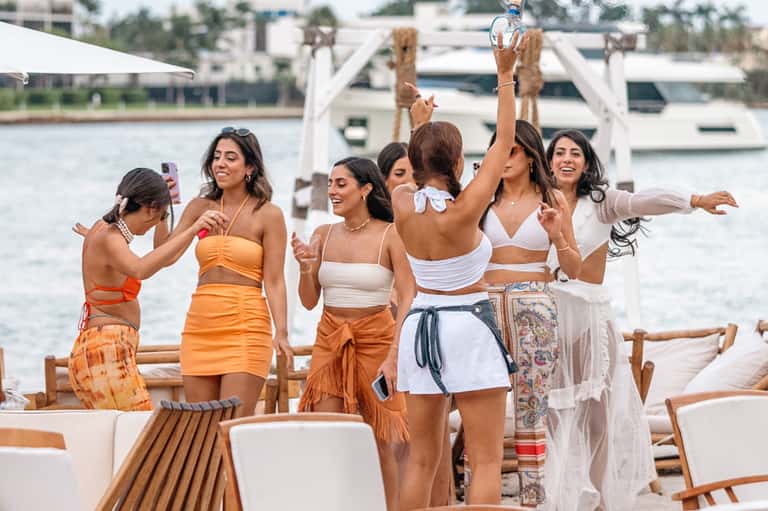 Women celebrating at waterfront deck with boats visible, holding drinks, enjoying social gathering
