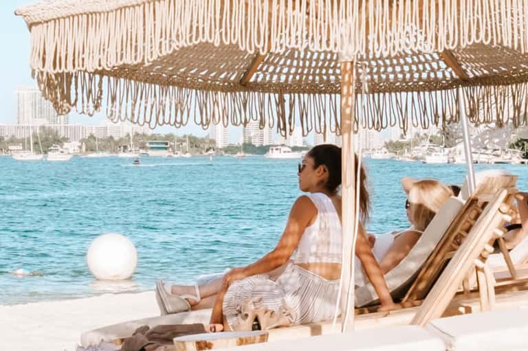 Beach lounge area with fringe umbrella, guests relaxing in chairs, ocean waterfront and coastal buildings visible
