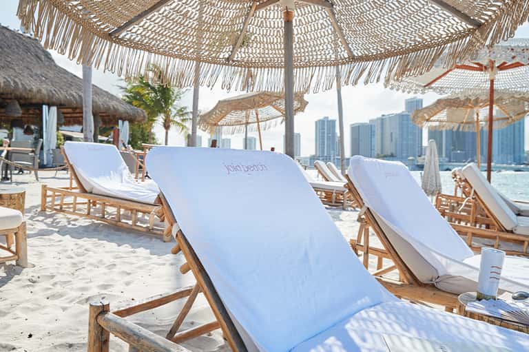 Beach club lounge with woven umbrellas, white loungers, wooden chairs, and city skyline backdrop