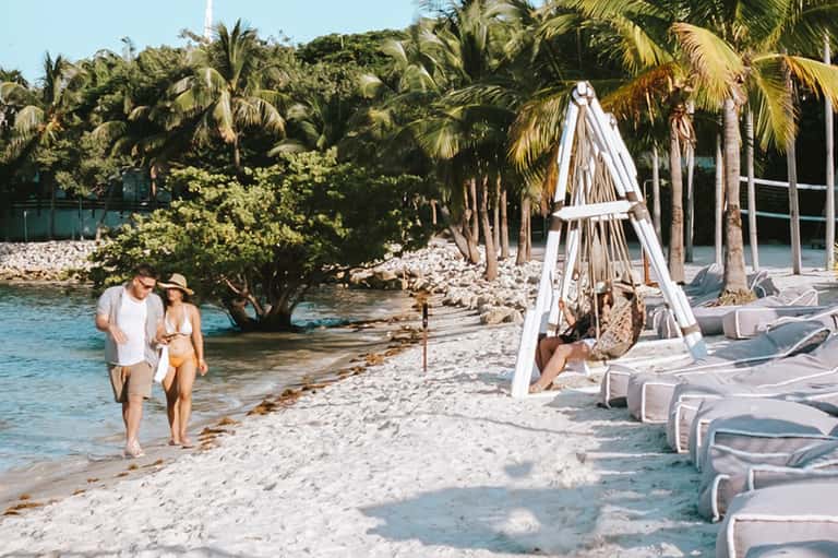 Couple walking along private beach with palm trees and white lounge chairs