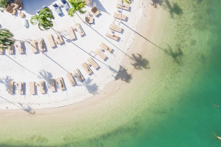 Aerial view of pristine white sand beach with lounge chairs and palm trees lining turquoise water
