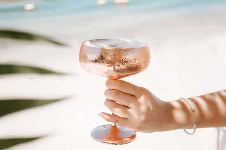 Hand holding rosé cocktail with frosted glass against blurred beach background