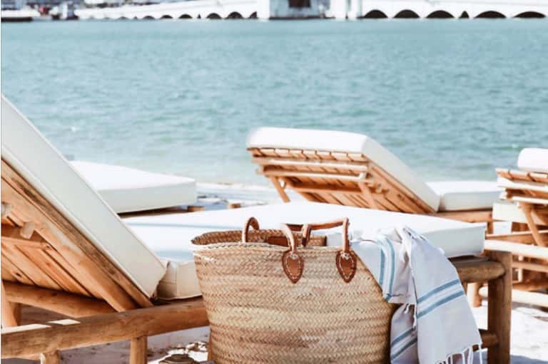 Wooden lounge chairs with white cushions and woven beach bag by ocean