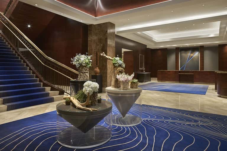 Luxury hotel lobby with blue-patterned carpet, curved staircase, and sculptural floral arrangements