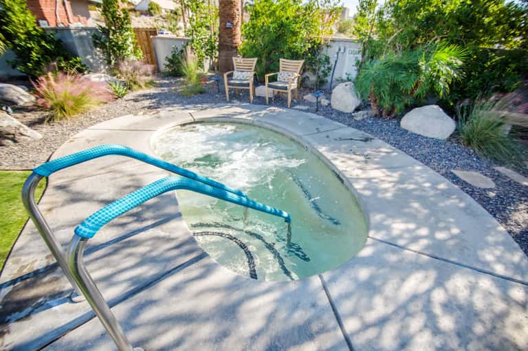 Neglected hot tub with algae and blue cleaning hose in landscaped backyard with palm trees