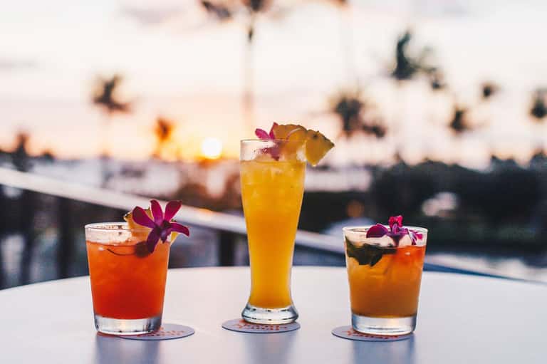 Three tropical cocktails with colorful flowers at oceanfront bar during golden hour