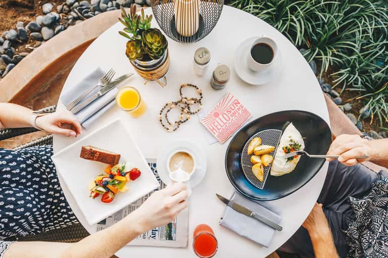 Outdoor café table setting with breakfast, coffee, juice, flowers, and two diners sharing meal