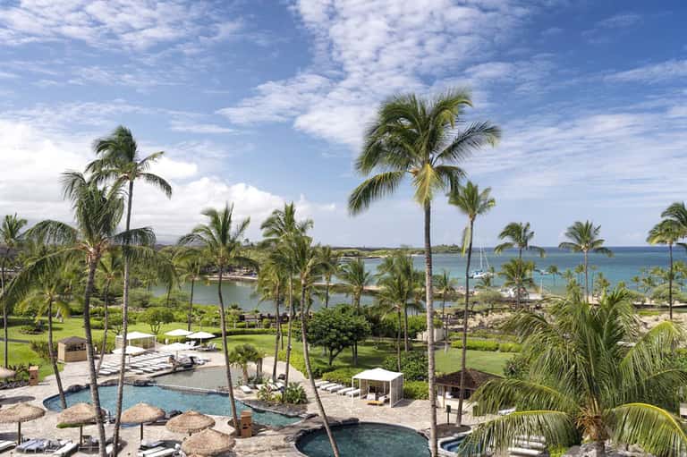 Aerial resort view with multiple pools, palm trees, manicured lawns, and ocean coastline in distance