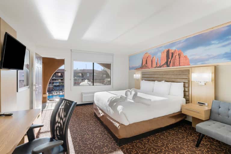 Modern hotel bedroom with queen bed, work desk, mountain artwork, and balcony with city views