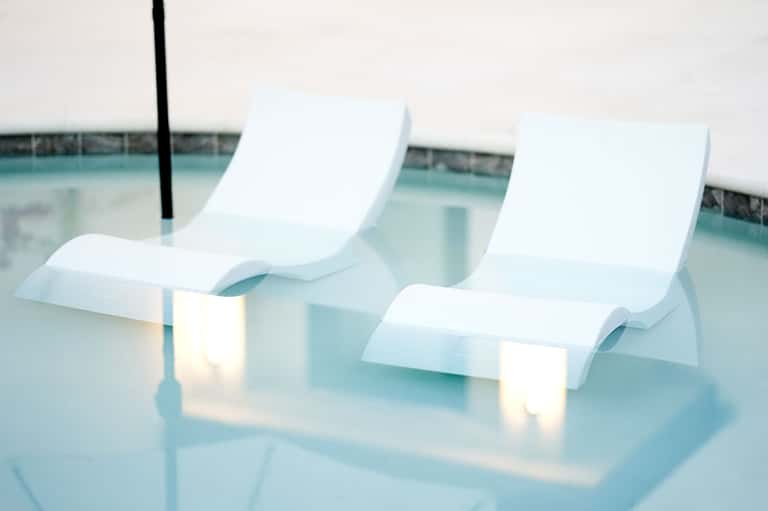 White curved lounge chairs sitting in shallow turquoise pool water with sun umbrella