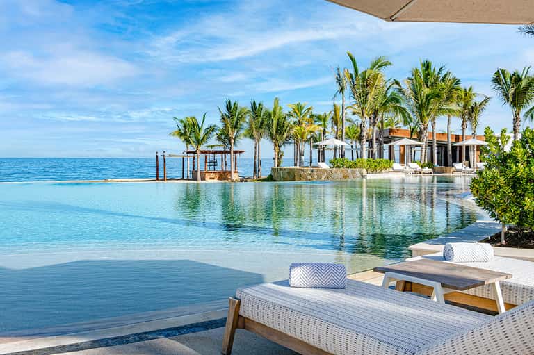Luxury beachfront infinity pool with palm trees, turquoise water, white lounge chairs, and ocean horizon view