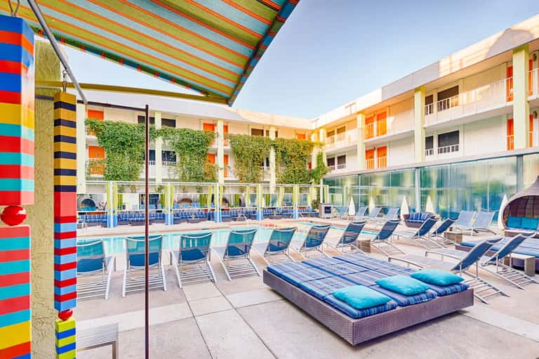 Colorful hotel courtyard with striped awning, lounge chairs, and modern residential buildings surrounding a sparkling pool.