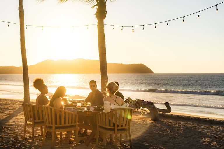 Group dining at beachfront table under string lights at sunset with ocean and palm trees