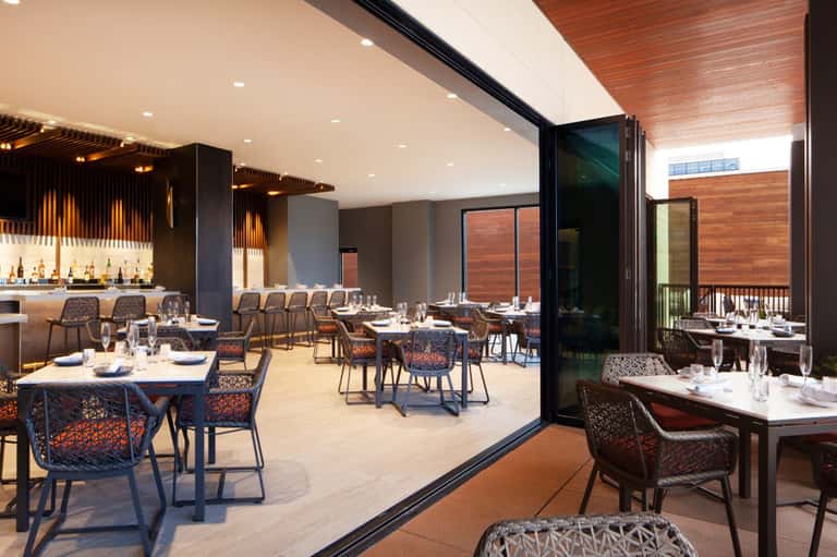 Modern restaurant with open-air design, wooden accents, elegant table settings, and architectural glass doors.