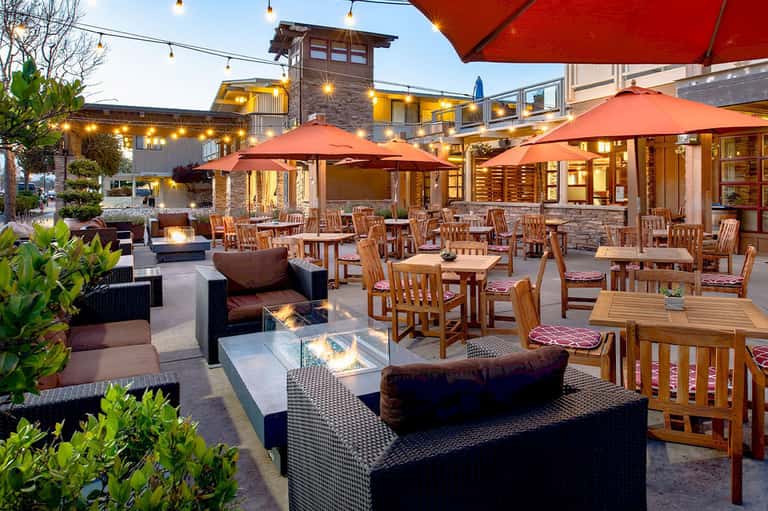 Outdoor terrace at dusk with wooden dining tables, red umbrellas, fire pit, and warm lighting