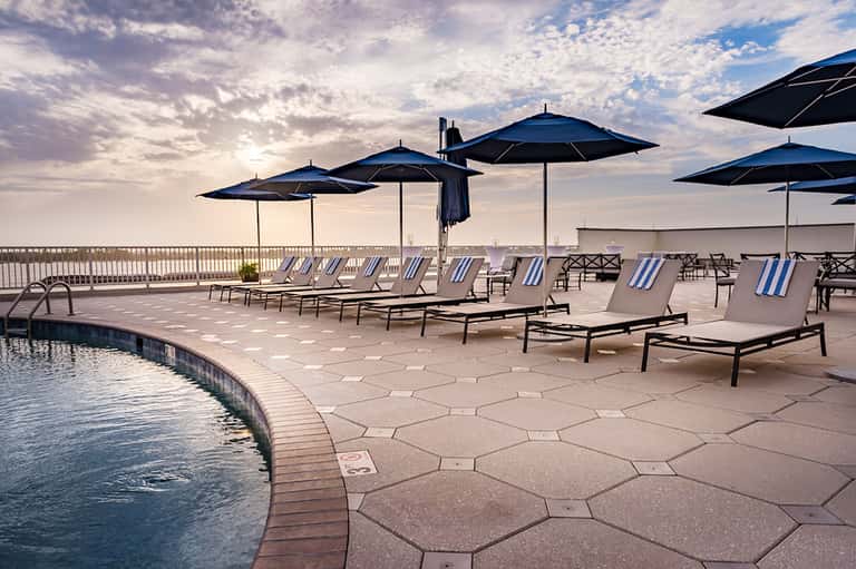 Rooftop pool deck with lounge chairs, blue umbrellas, ocean view, and evening sky