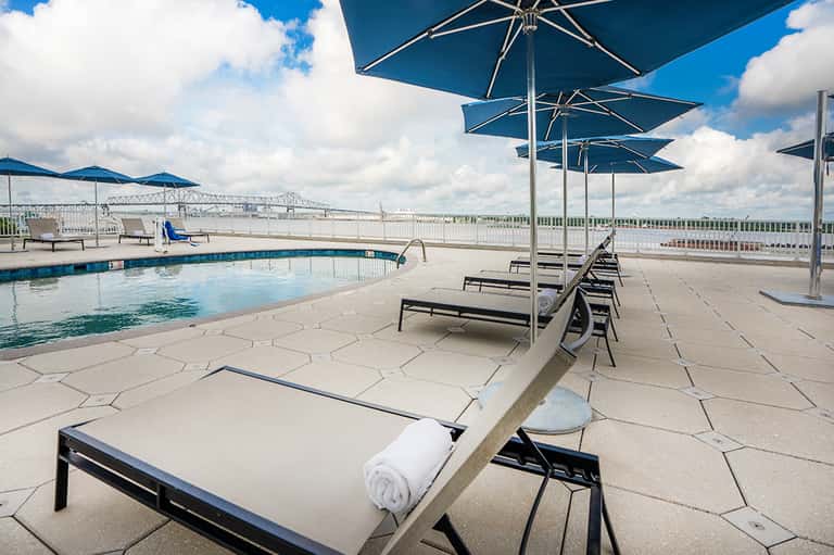 Rooftop pool with blue umbrellas, loungers, river view, and architectural bridge in background