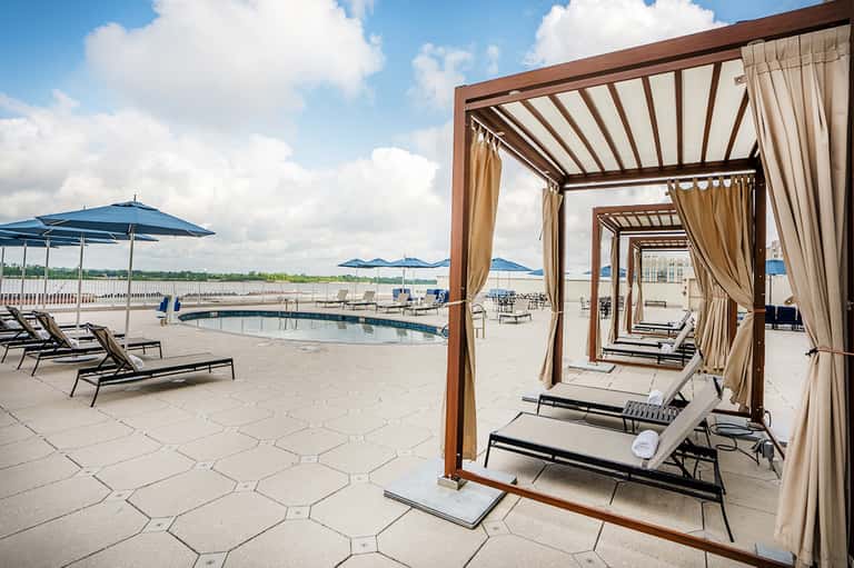 Waterfront pool deck with brown pergolas, tan curtains, blue umbrellas, and water view