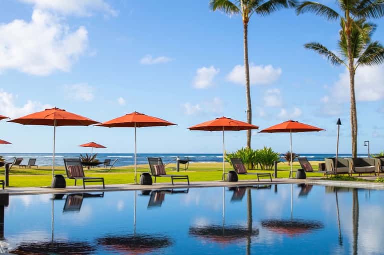 Resort pool area with orange umbrellas, lounge chairs, palm trees, manicured lawn, and ocean horizon