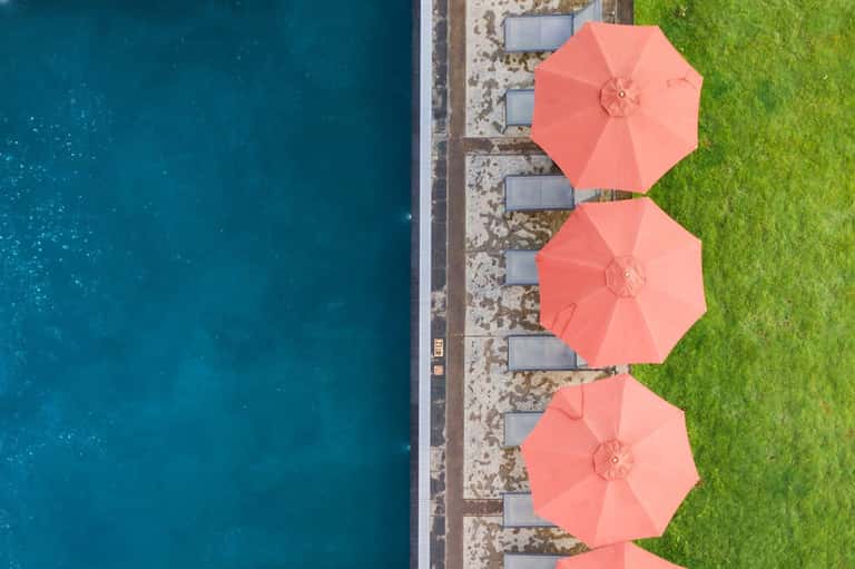 Aerial view of three coral umbrellas on manicured lawn beside dark blue water and white pier