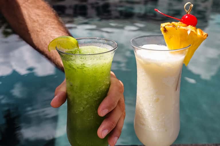 Two tropical cocktails held by person near pool, lime-colored and pineapple drinks