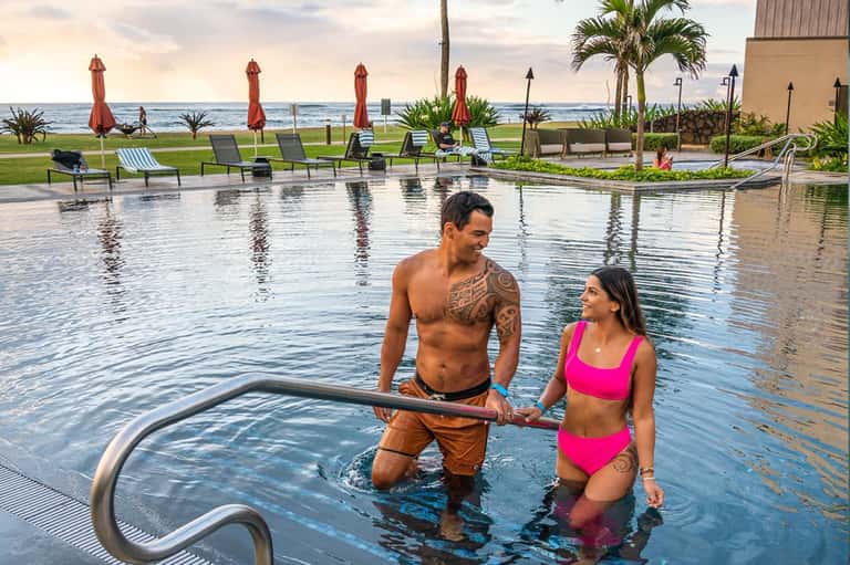 Couple in pink swimwear in infinity pool overlooking ocean at dusk with red umbrellas