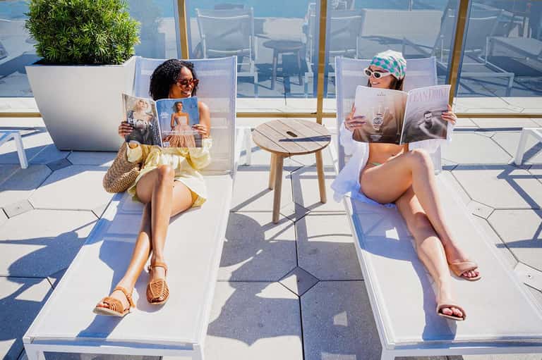Women reading magazines on cushioned lounge chairs on rooftop terrace overlooking ocean and sky