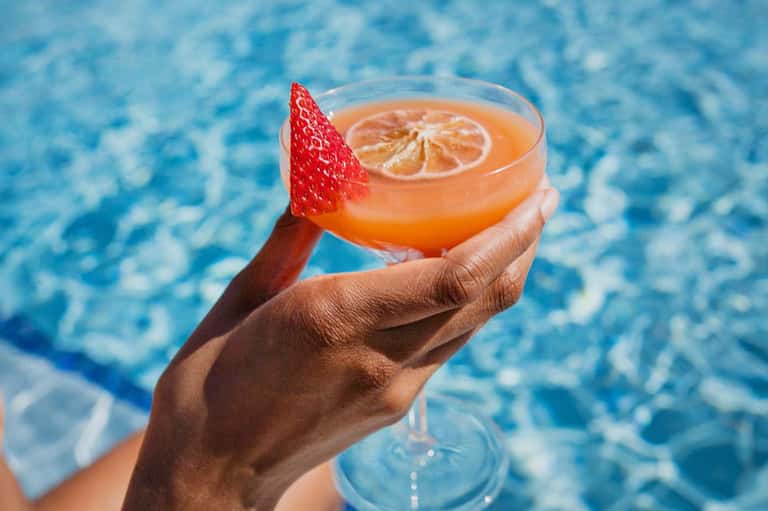 Hand holding refreshing tropical cocktail with strawberry and citrus slice over pool water