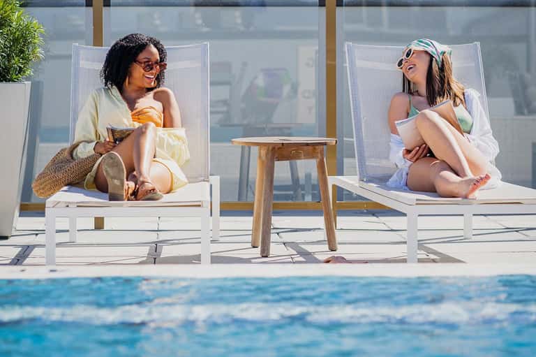 Two women relaxing on white lounge chairs by pool with ocean view and glass wind barriers