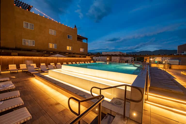 Illuminated rooftop pool at dusk with lounge chairs and mountain views