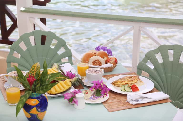 Beachfront breakfast table with tropical flowers, fresh pastries, juice, and ocean view beyond railing