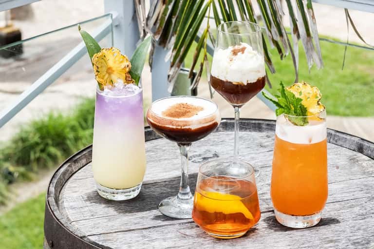 Four specialty cocktails with tropical garnishes displayed on a black wooden table outdoors