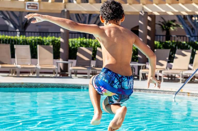 Child about to jump into a clear blue swimming pool with lounge chairs visible in background