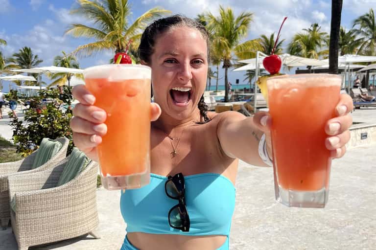 Woman holding two colorful tropical drinks at a beach bar with palm trees in background