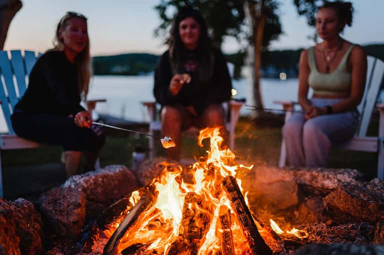 Group of people roasting marshmallows around a campfire by a lakeside at dusk