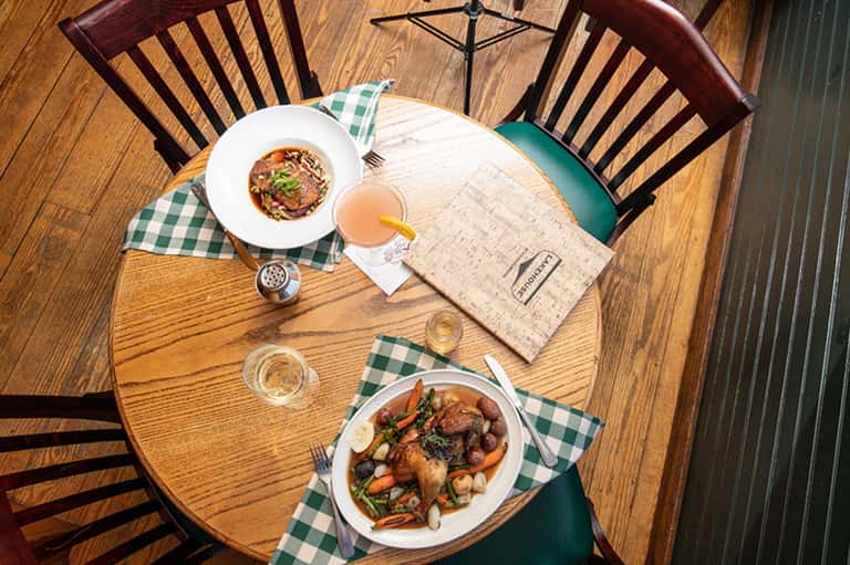 Overhead view of dining table with gourmet plated dishes, beverage, and green gingham napkins on wood surface