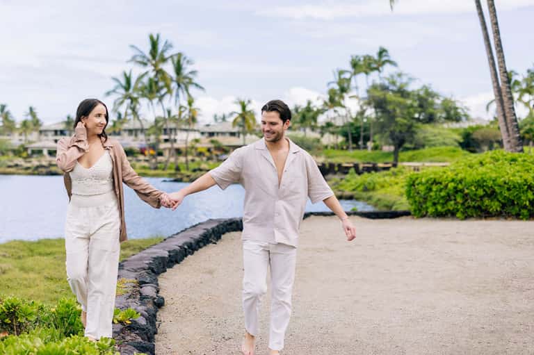 Couple holding hands walking on beach with palm trees and resort in background