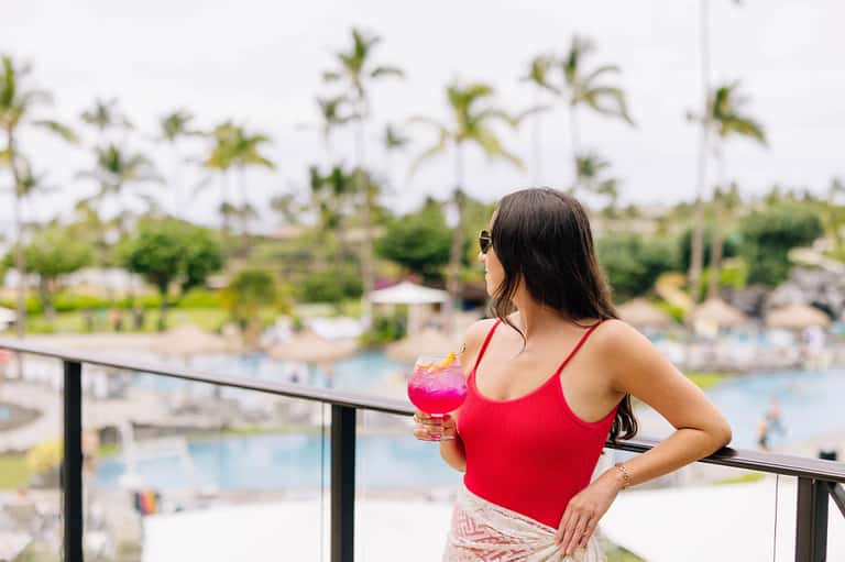 Woman on balcony holding pink cocktail overlooking resort pools and tropical landscaping below