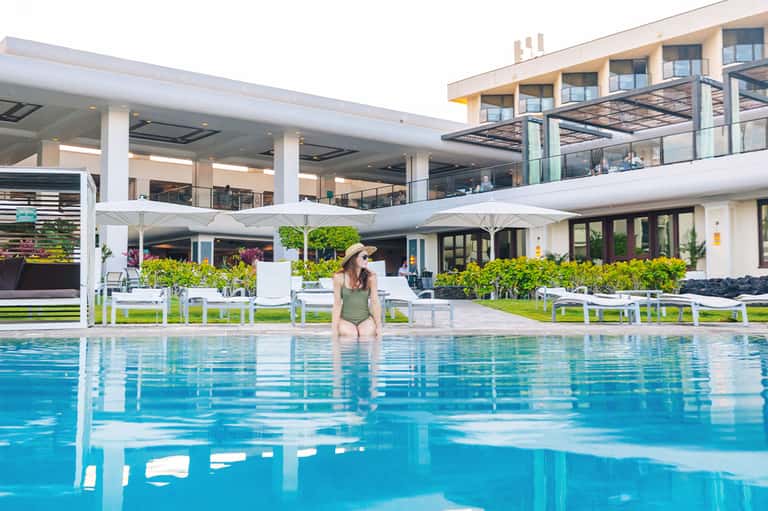 Woman in green swimsuit wading in resort swimming pool with white umbrellas and building